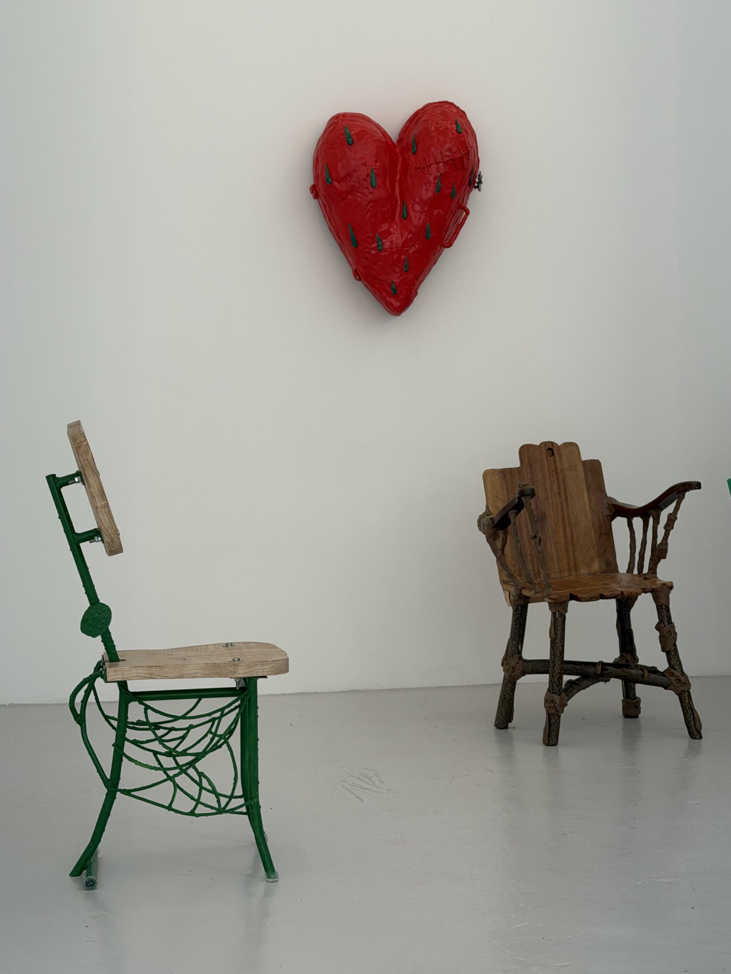 This image captures an intriguing indoor display featuring a vibrant red heart sculpture on a white wall, flanked by two distinctively crafted chairs